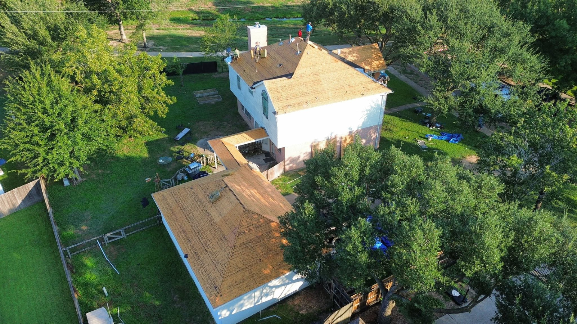 Aerial view of a two-story home with its roof stripped to bare wooden decking while roofers work on the roof