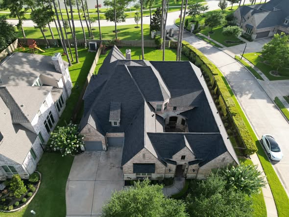 Drone view of a large suburban home with a dark architectural shingle roof, surrounded by pine trees