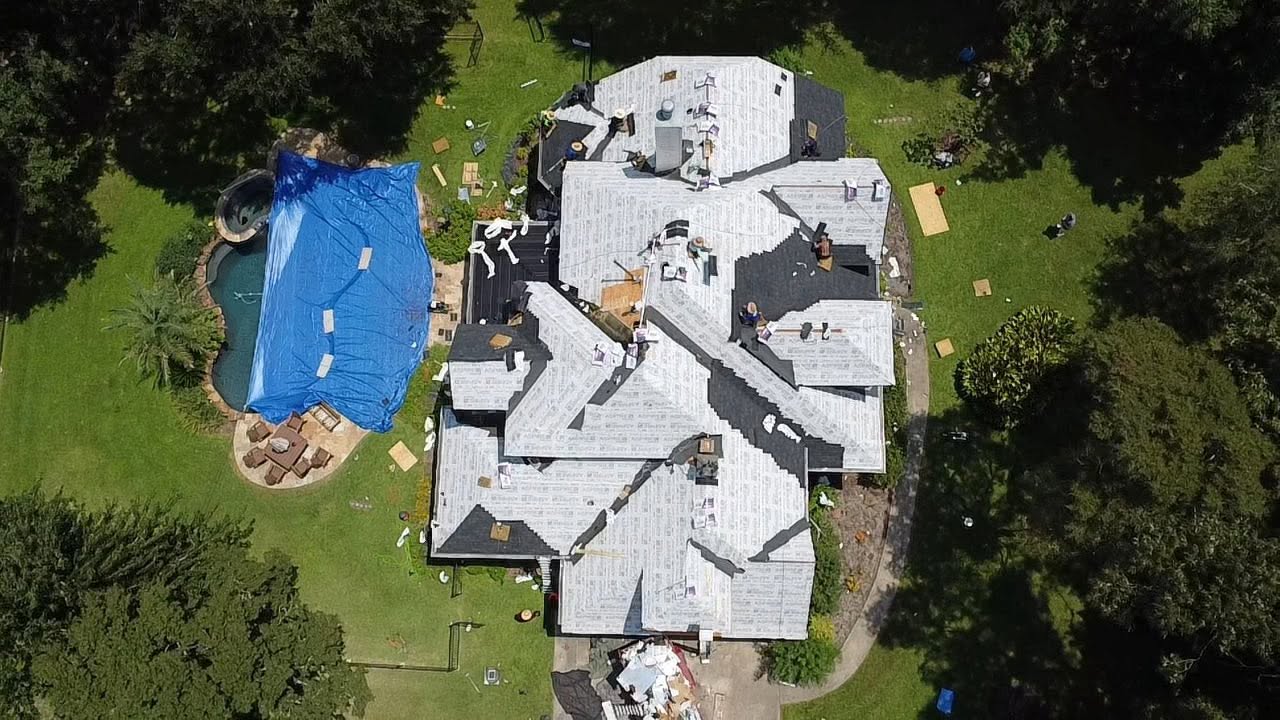 Aerial view of a crew replacing a roof on a large home, with underlayment and new dark shingles on the roof and a covered pool in the backyard