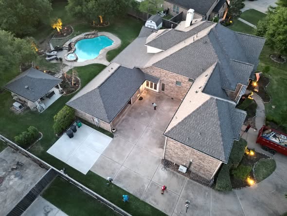 Drone view of a large brick home with a dark shingle roof, a debris trailer and crew on the driveway