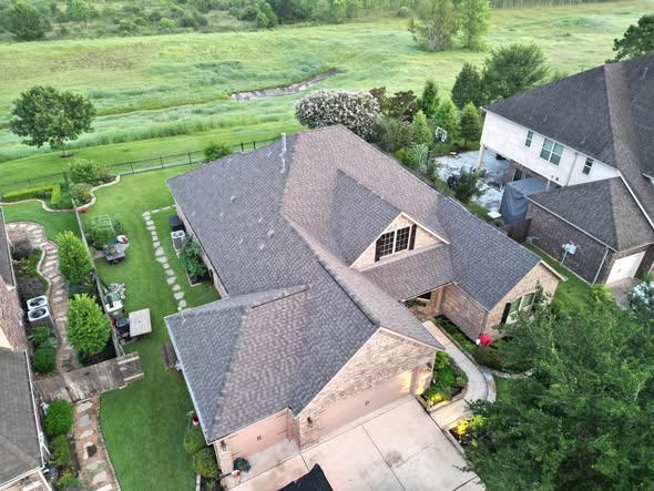 Drone view of a two-story brick home with a multi-gable brown and grey shingle roof